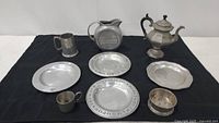 Full set of pewter serving pieces arranged on black cloth, including coffee urn, pitcher, four plates, mug, cup, and small round dish.