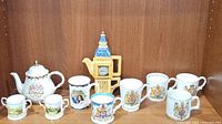 Front view of all items together: white ceramic teapot, Big Ben teapot, and 8 commemorative cups.
