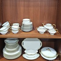 Front view of entire Theodore Haviland Limoges China set including cups, saucers, teapot, cream pitcher, sugar dish, casserole, and platter arranged on wooden shelf.