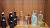 Fourteen vintage bottles arranged on a wood shelf including beer/soda, milk, and medicine bottles.