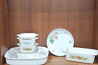Vintage Corning Ware set with five casserole dishes stacked and arranged around a ceramic pie plate inside a wood cabinet.