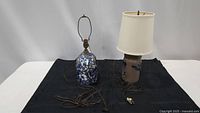 Overview of two table lamps placed on black cloth, showing the blue and white crackled ceramic lamp without shade and the brown stoneware lamp with a beige shade and power cords extended