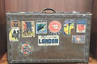 Front view of the vintage suitcase showing travel stickers from various locations and the handle with leather tag.