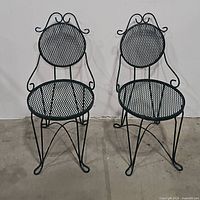Two black wrought iron mesh bistro chairs with round seats and backs, ornamental scrollwork on the backrest, and curled feet on the legs.