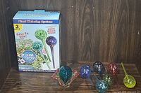 Full view of six colorful hand blown glass plant watering globes arranged next to a box and a decorative hand blown glass dish with ruffled edges.