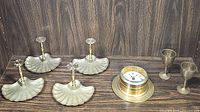 Four brass shell curtain pull backs, nautical clock, and two brass wine glasses displayed on wooden surface.