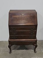 Full front view of vintage wooden bureau desk with closed slant top and four drawer fronts with metal pulls, showing antique wood finish and carved legs.