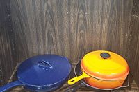 Blue enameled round covered pot and orange to yellow gradient enameled skillet with lid on wooden shelf