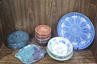 Photo showing the full lot - blue and white decorative plate, dark blue lidded bowl, green stoneware dish, stacked small brown bowls, and three small blue/white glazed bowls