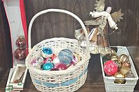 A white wicker basket filled with colorful vintage and plastic Christmas balls including blue, pink, red, white, and silver shiny and matte ornaments along with a large angel figurine standing behind baskets containing more ornaments.