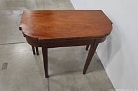 Folded view of rectangular mahogany flip-top table with rounded edges and four straight legs on concrete floor against white wall.
