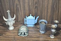 Photo showing all six metal decorative items together on a wooden surface: tribal statue, turtle figurine, teapot wall decor, metal cup, and two vases.