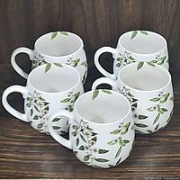 Five white Konitz porcelain snuggle mugs with green leaf and white flower pattern arranged in a group on a wooden surface.