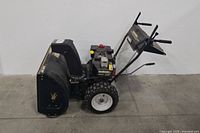Full side view of the black snow thrower showing the large tires, chute, and control handles.