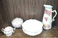 Four vintage porcelain items displayed against wood background: pink floral bowl and plate, footed lidded dish, sugar dish, and floral pitcher.