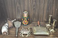 Overview photo showing all items in the lot on a wooden surface: horn flask with strap, metal hand brush, Russian nesting doll, crab inkwell, silent butler, and candlestick holders.