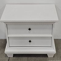 Front view of a white nightstand with two closed drawers showing simple black knobs and a base wider than the main body.