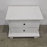 Front view of white nightstand with closed drawers showing overall shape, color, and hardware.