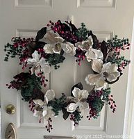 Christmas wreath hanging on a white door, showing front view with white flowers, red berries, and green foliage decoration.