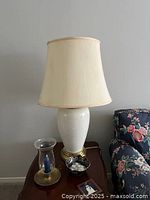 Full view of cream ceramic lamp standing on table beside floral armchair, showing lamp base and fabric shade.
