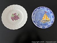 Two decorative plates side by side on a dark surface: left is white textured plate with pink bougainvillea flowers, right is blue plate with yellow sailboat design.