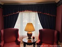 Wide shot of window with navy blue drapes, cranberry trimmed valance and side panels hung with tiebacks, highlighting the elegant setup in a formal room with wing chairs and table lamp.