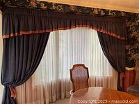 Full view of formal navy blue draperies with a cranberry trim valance, sheer white lining behind. Draperies are tied back with cranberry tiebacks beside a round dining table and cane back chair.