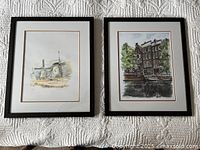 Two framed watercolor prints from Amsterdam, showing a windmill and Anne Frank's House, laid flat on white quilted fabric.