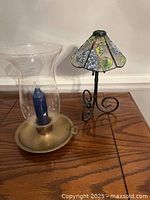 Two candle holders side by side, one a clear glass hurricane with blue candle, the other a colorful stained glass tea light holder on a black metal stand.