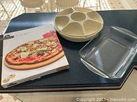 All three items shown together on countertop: boxed Grillpro pizza stone, Anchor Hocking glass casserole dish, and Tupperware veggie tray with lid.