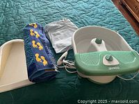 Conair foot spa, heating pad, and towel lying on quilted teal fabric surface. Spa is white with mint green accents, control knob on front.