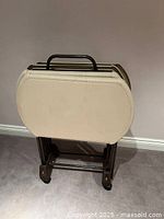 Four cream-colored plastic TV trays nested together on a brown metal trolley with castor wheels and handle, viewed from side against beige wall.