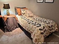 Full view of twin bed with bedding set showing duvet cover in multicolor leaf pattern and 4 pillows arranged on bed, bedside table with brown cover and lamp beside bed.
