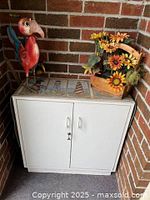White two-door laminate cabinet with decorative runner and decorative items on top, placed against brick wall.