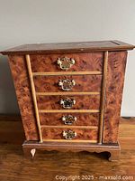 Front view of closed wooden jewellery box showing five drawers with decorative brass handles and burl wood veneer finish.
