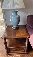 Solid hardwood wooden end table with a light-colored ceramic vase lamp on top, showing patterned wood grain on the tabletop and the drawer below.
