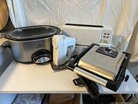 Four kitchen appliances on white table: Rival slow cooker, KitchenAid hand mixer, KitchenAid toaster, Bella panini press
