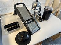 Overview of the mandoline slicer with five blade attachments, hand guard, French press, and electric coffee grinder on a white table.