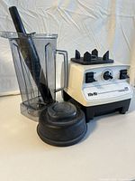 Photo of Vita-Mix 5000 blender with container, black tamper, and lid sitting next to the motor base on white surface with white background.