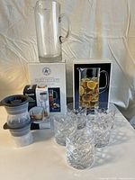 Full view of the lot showing 2 glass pitchers on boxes, 5 cut pattern highball glasses in front, and 4 Tovolo sphere ice molds stacked to the side. White tablecloth and neutral backdrop.