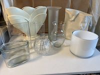 Nine glass and ceramic vases arranged on white surface against beige background.