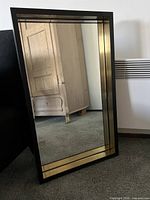 Front view of rectangular Crate & Barrel mirror with black frame and gold inner trim, standing on carpet.