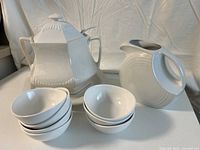 Photo of white ceramic hexagonal soup tureen with lid, underplate, six small bowls, and a pitcher on a white surface with beige backdrop.