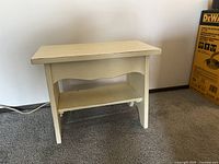 Front view of a small wooden bench painted off-white with a lower shelf against a white wall and carpeted floor.