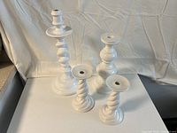 Four white ceramic candle holders shown on white table against cloth backdrop, varying heights and designs visible