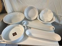 Lot of six white ceramic serving dishes including oval, round, scalloped bowls, and long trays displayed on a white tray table
