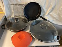 Photo of Tramontina paella pan (with glass lid), Scanpan frying pan (with glass lid), Nordic Ware frying pan (no lid), orange silicone steamer/warmer, and metal wire cooling/baking rack on white background.