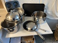 Overview of entire lot showing all cookware including pot with steamer insert and domed lid, frying pans, muffin tin, and roasting tray