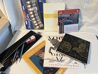 Art supplies displayed on white surface with acrylic paint set, brushes, coloring books, sketch pads, pencil case, and pencil