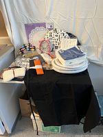 Photo showing assortment of kitchen textiles including black apron, tea cozys with various patterns, paper doilies, paper plates, and pot holders laid out on table.
